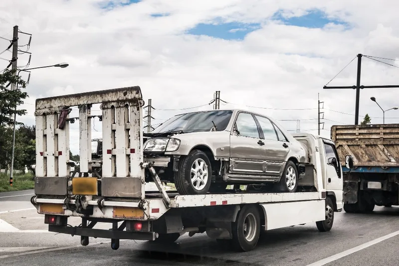Damaged sedan loaded on a small flatbed truck beside a scrap hauler